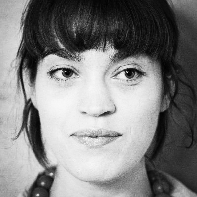 A light-skinned woman with a dark choppy fringe and well-defined eyebrows looks sideways at the camera. She is in front of a plain grey background and her face fills the picture.