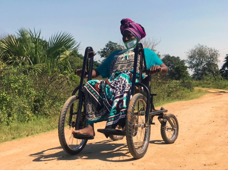 A local Kenyan lady with a bright purple turban and teal patterned dress drives her Safariseat at speed down a dusty track, leaving a cloud of golden dust in the air behind her.