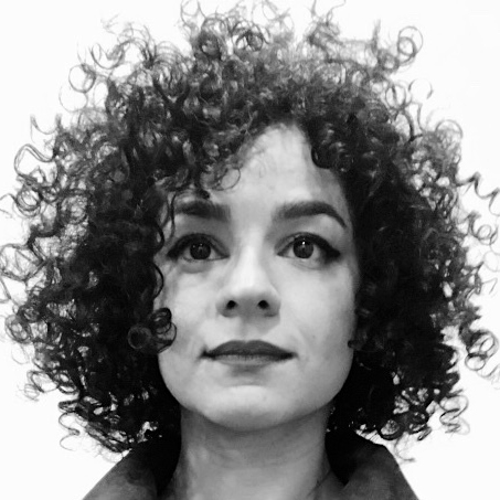 A light-skinned person with dark tightly curled hair is looking upwards and smiling slightly. She has wide open dark eyes and is stood in front of a white background.