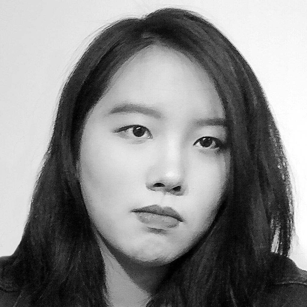 A light-skinned young woman with dark side-parted hair staring seriously. She is standing in front of a white background and her head is slightly tilted.